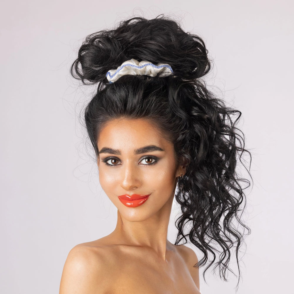 A dark-haired smiling woman, front view, with stylish ponytail is wearing Malkiele designer white silk scrunchie, embellished light blue silk knit ribbon, name Morning Passion.