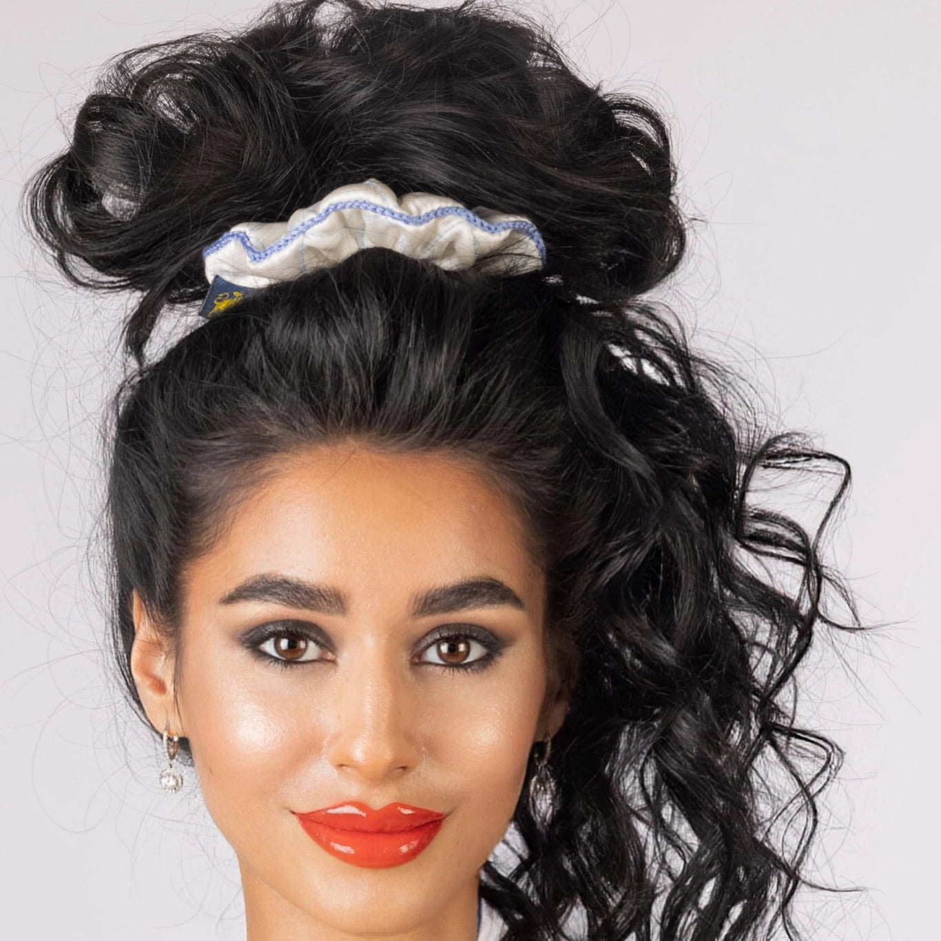 
                  
                    A dark-haired smiling woman, front view, with stylish ponytail is wearing Malkiele designer white silk scrunchie, embellished light blue silk knit ribbon, name Morning Passion.
                  
                