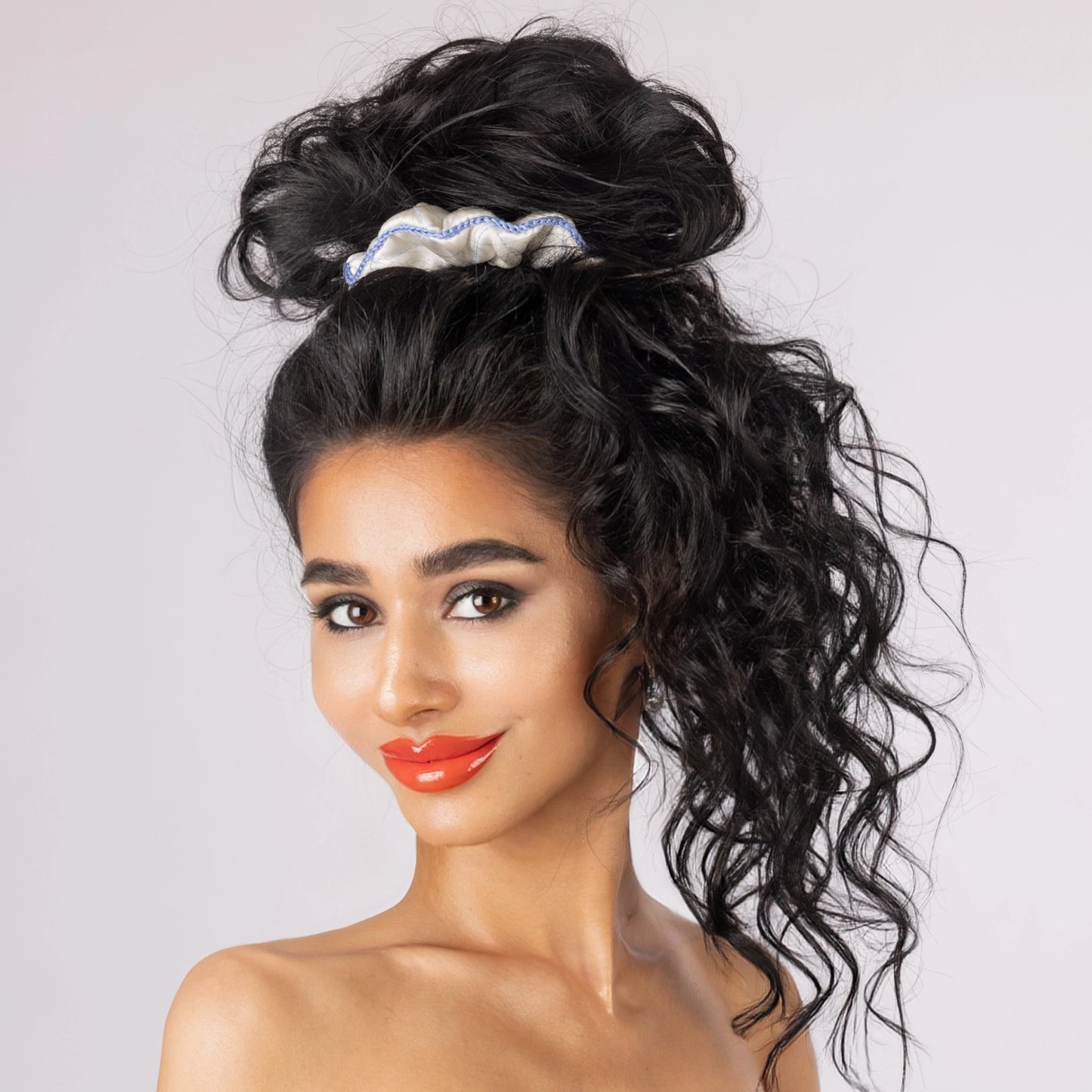 
                  
                    A dark-haired smiling woman, front view, with stylish ponytail is wearing Malkiele designer white silk scrunchie, embellished light blue silk knit ribbon, name Morning Passion.
                  
                