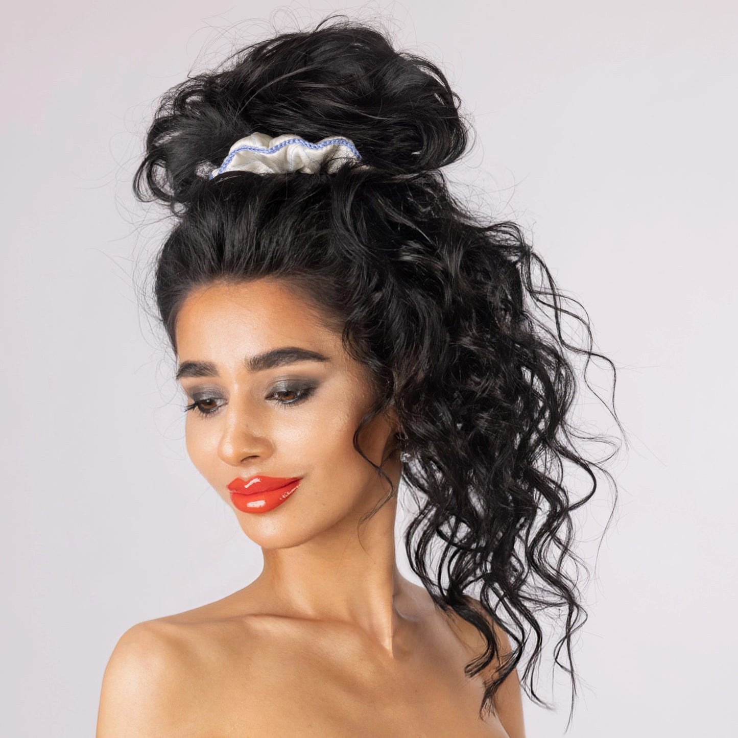 
                  
                    A dark-haired smiling woman, front view, with stylish ponytail is wearing Malkiele designer white silk scrunchie, embellished light blue silk knit ribbon, name Morning Passion.
                  
                