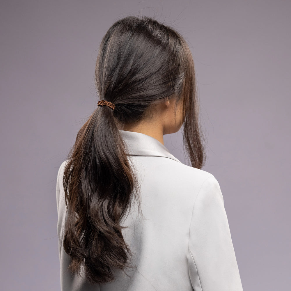 
                  
                    A dark-haired woman in grey jacket, view from the back, with a stylish ponytail is wearing Malkiele designer brown silk hair tie, name Finesse.
                  
                