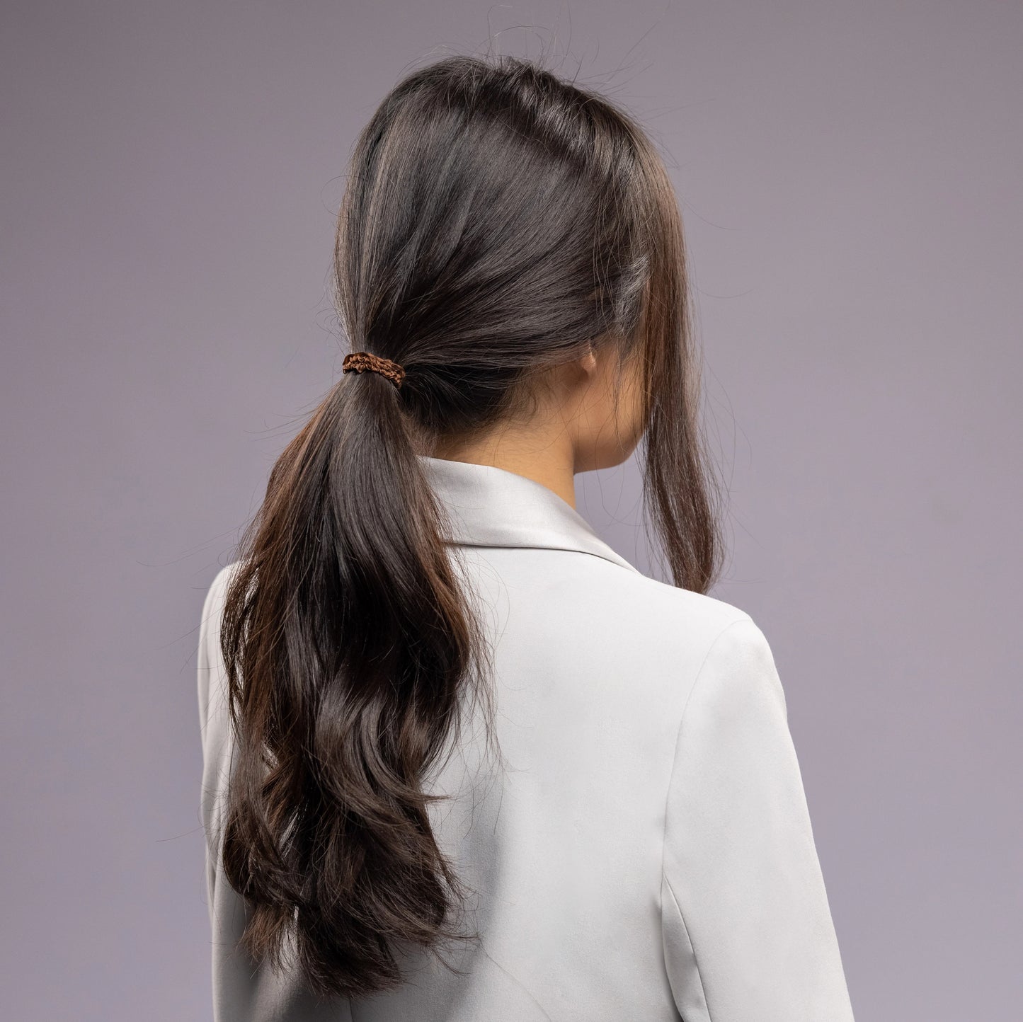 A dark-haired woman in grey jacket, view from the back, with a stylish ponytail is wearing Malkiele designer brown silk hair tie, name Finesse.