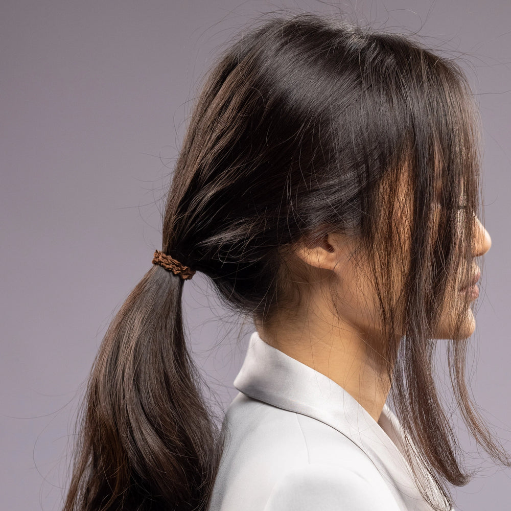 
                  
                    A dark-haired woman in grey jacket, side view with half face, with a stylish ponytail is wearing Malkiele designer brown silk hair tie, name Finesse.
                  
                