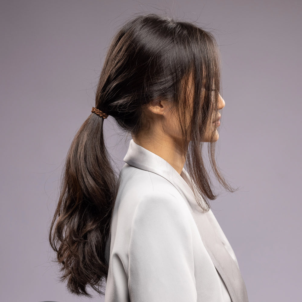 
                  
                    A dark-haired woman in grey jacket, side view with half face, with a stylish ponytail is wearing Malkiele designer brown silk hair tie, name Finesse.
                  
                