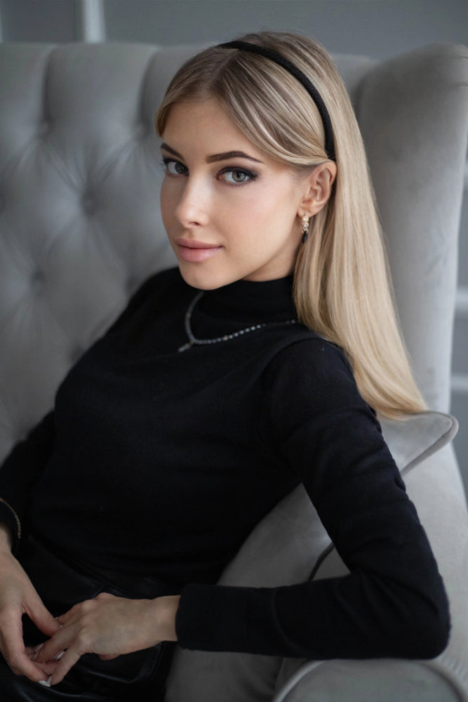 
                  
                    A blonde-haired girl in black turtleneck, looks at the camera, with flowing hair is wearing Malkiele designer black silk knit headband, name Virgin.
                  
                