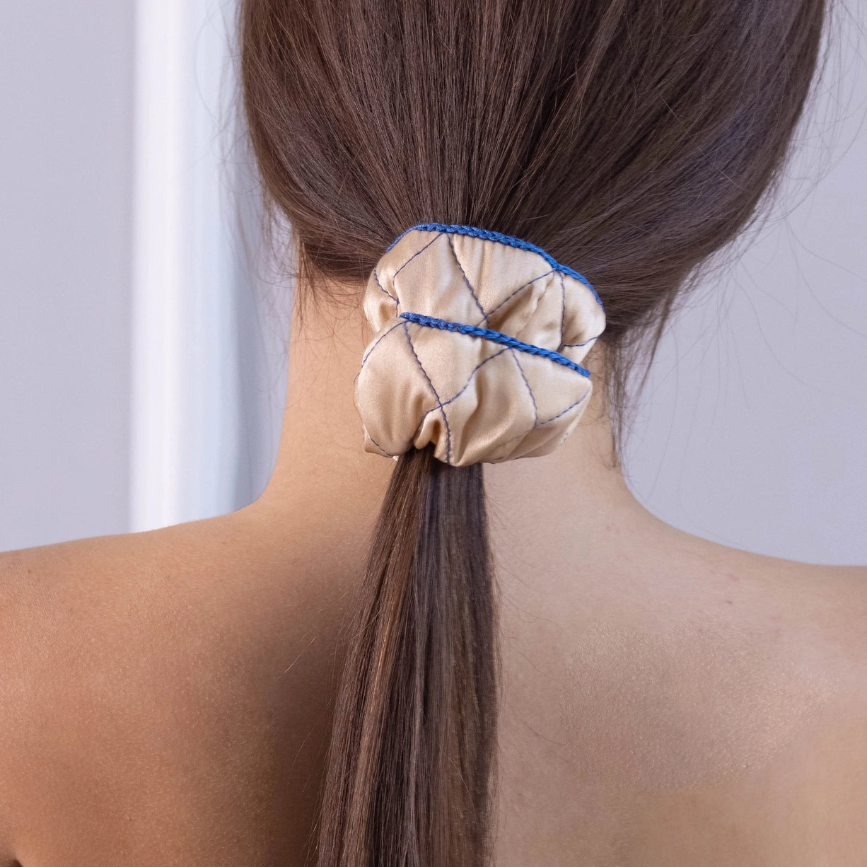 
                  
                    A dark-haired woman, view from the back, with stylish ponytail is wearing Malkiele designer beige silk scrunchie, embellished blue silk knit ribbon, name Morning Passion.
                  
                