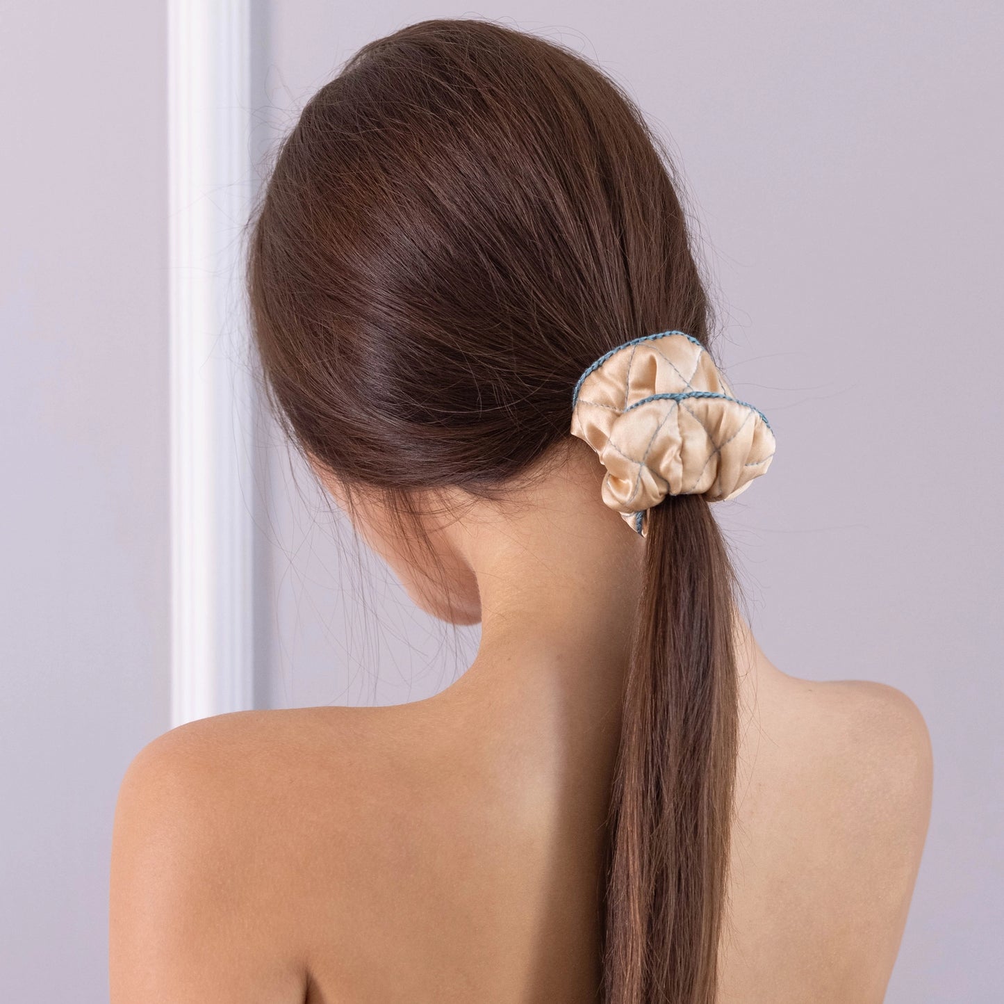 
                  
                    A dark-haired woman, view from the back, with stylish ponytail is wearing Malkiele designer beige silk scrunchie, embellished green silk knit ribbon, name Morning Passion.
                  
                