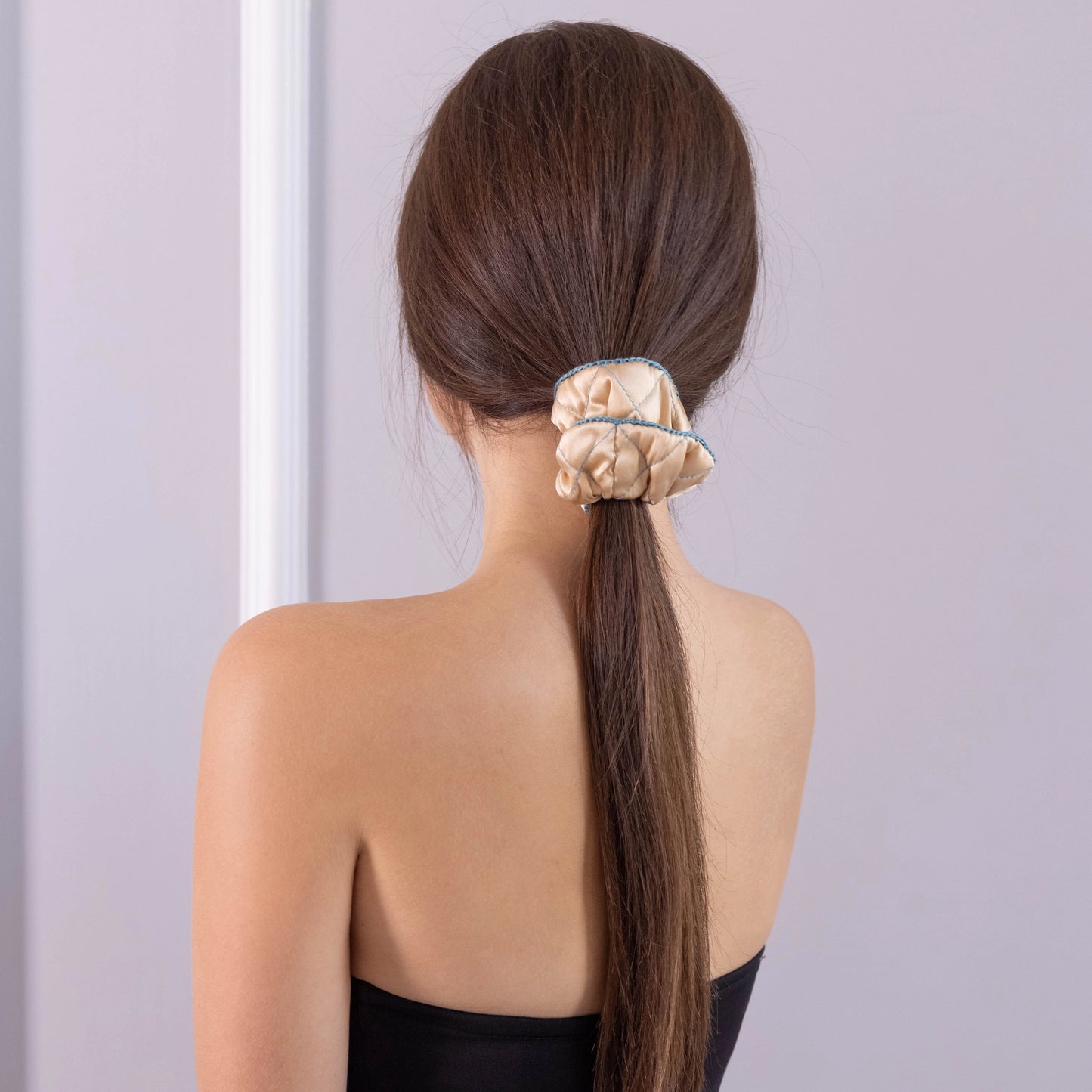 A dark-haired woman, view from the back, with stylish ponytail is wearing Malkiele designer beige silk scrunchie, embellished green silk knit ribbon, name Morning Passion.