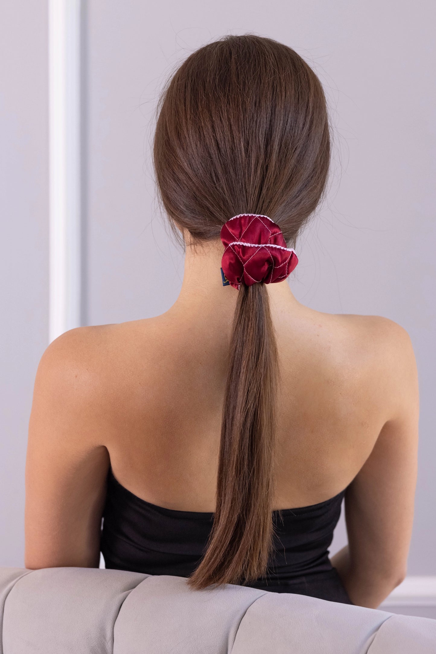 
                  
                    A dark-haired woman, view from the back, with stylish ponytail is wearing Malkiele designer burgundy silk scrunchie, embellished pink silk knit ribbon, name Morning Passion.
                  
                