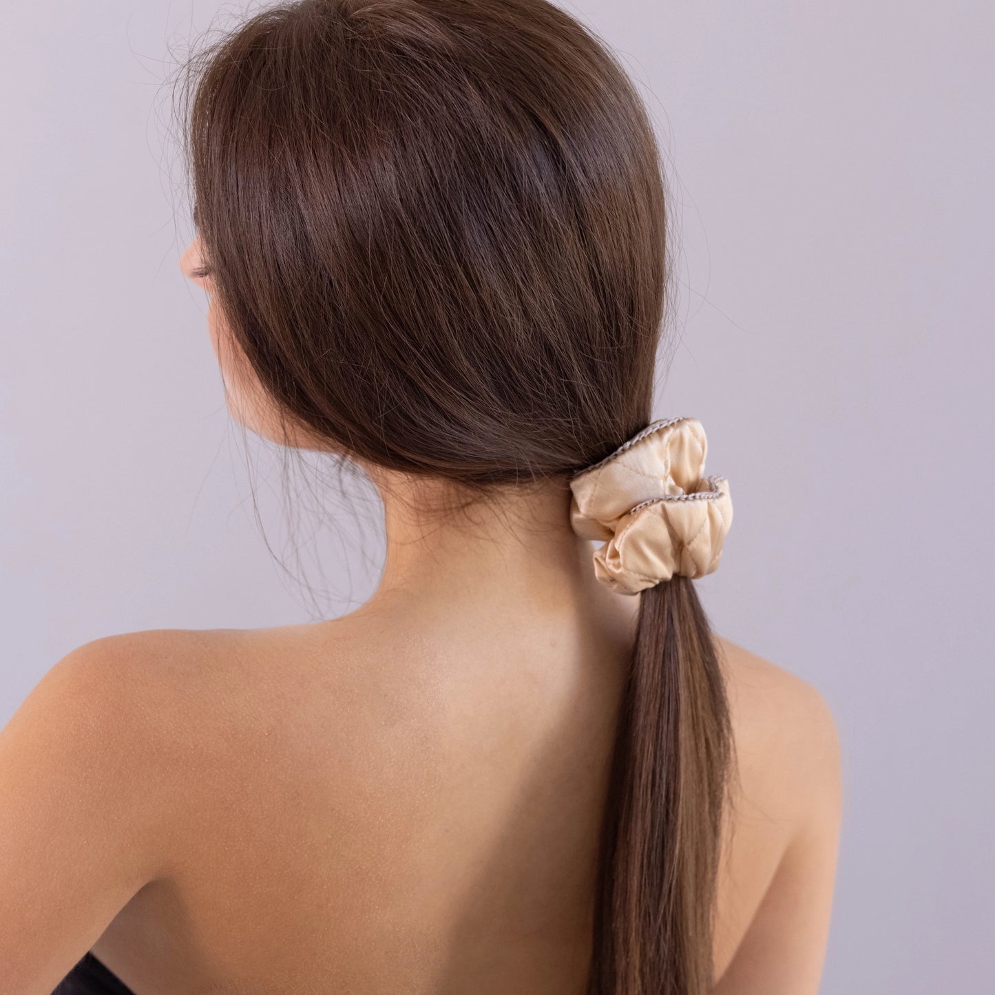 A dark-haired woman, view from the back, with long and stylish ponytail is wearing Malkiele designer beige silk quilted scrunchie, embellished with beige silk knit ribbon, name Morning Passion.