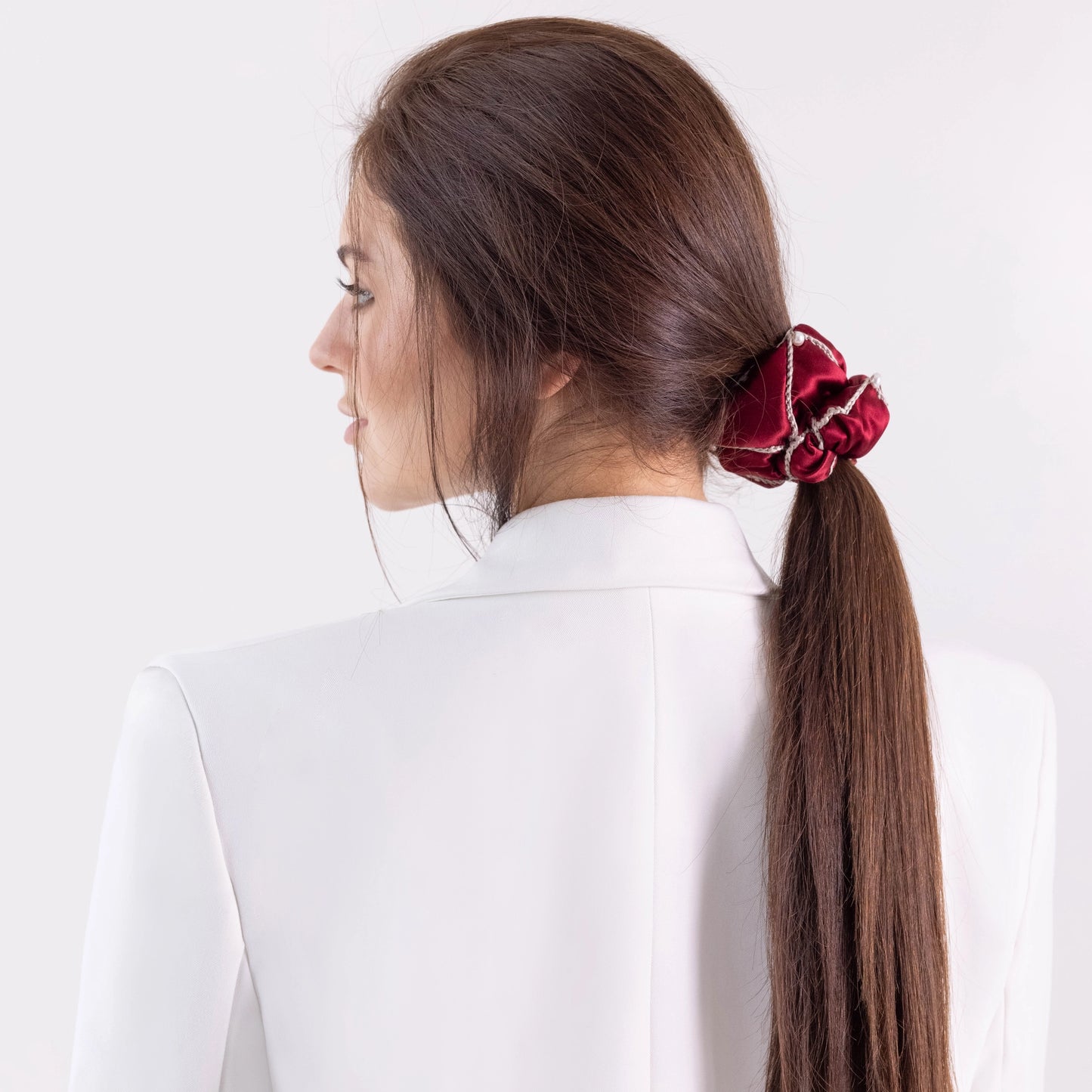 Dark-haired woman in white jacket, with stylish ponytail is wearing Malkiele designer burgundy silk scrunchie, embellished with 6 white pearls from Swarovski® and beige silk knit ribbon, name Majestic.