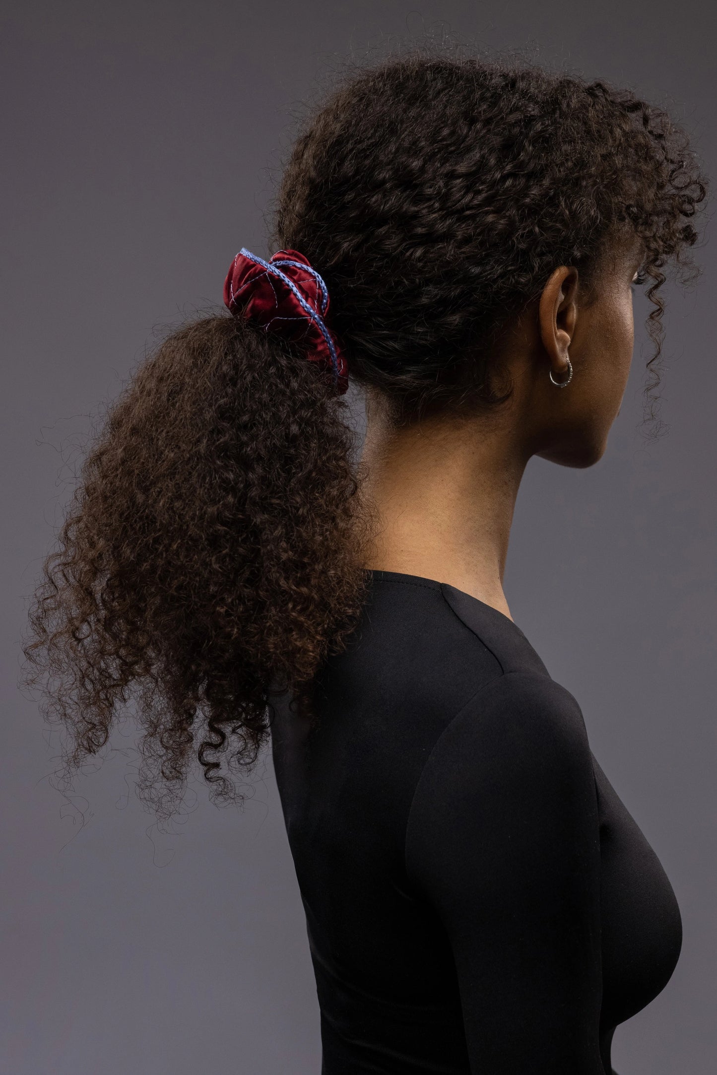 
                  
                    An African woman, view from the side, with stylish ponytail is wearing Malkiele designer burgundy quilted silk scrunchie, embellished with light blue silk knit ribbon, name Morning Passion
                  
                