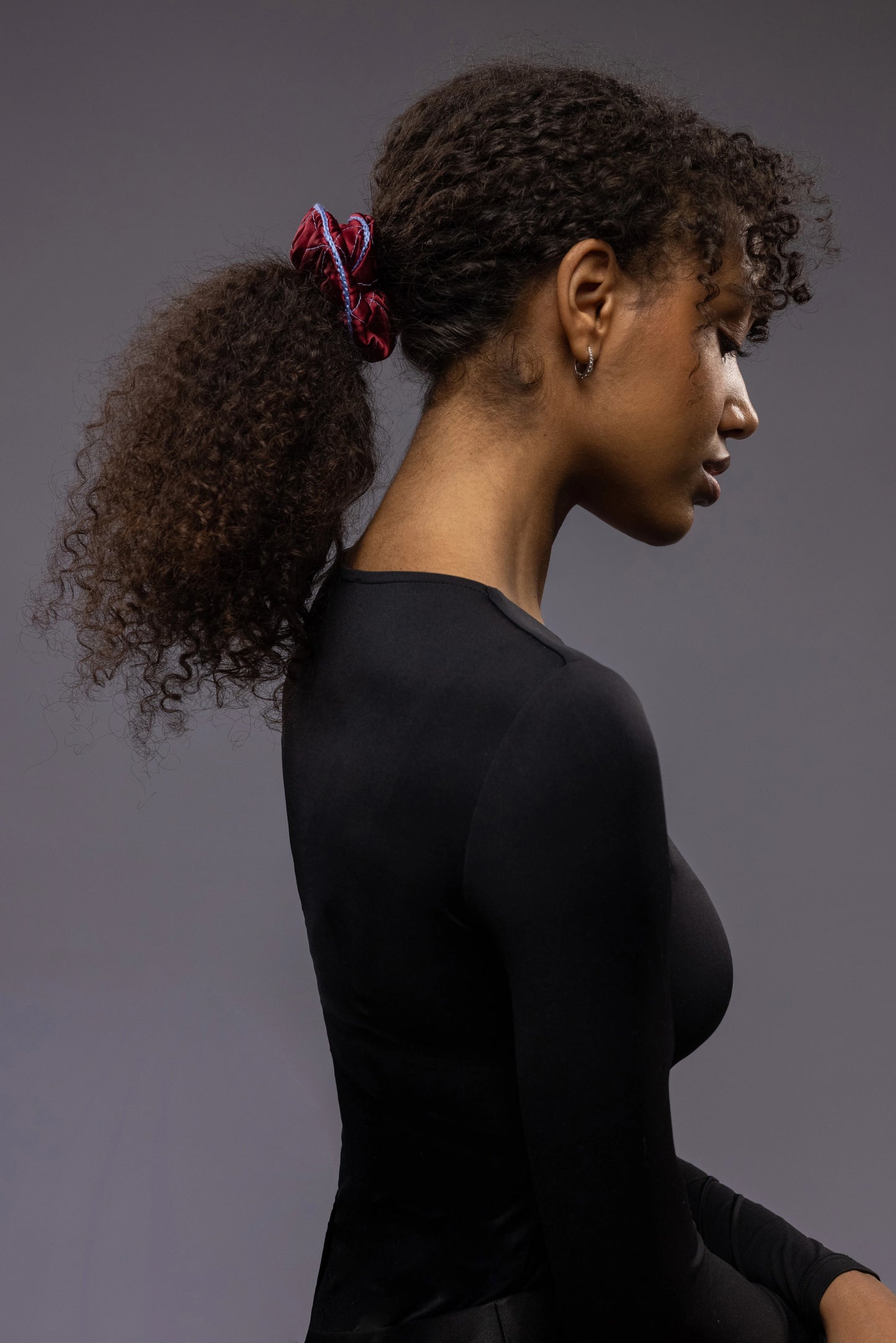 
                  
                    An African woman, view from the side, with stylish ponytail is wearing Malkiele designer burgundy quilted silk scrunchie, embellished with light blue silk knit ribbon, name Morning Passion
                  
                