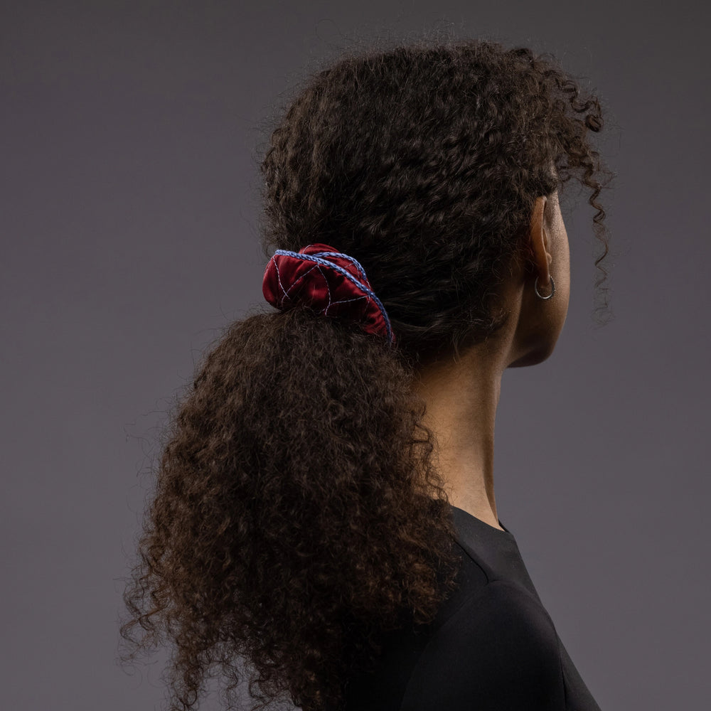 
                  
                    An African woman, view from the side, with stylish ponytail is wearing Malkiele designer burgundy quilted silk scrunchie, embellished with light blue silk knit ribbon, name Morning Passion
                  
                