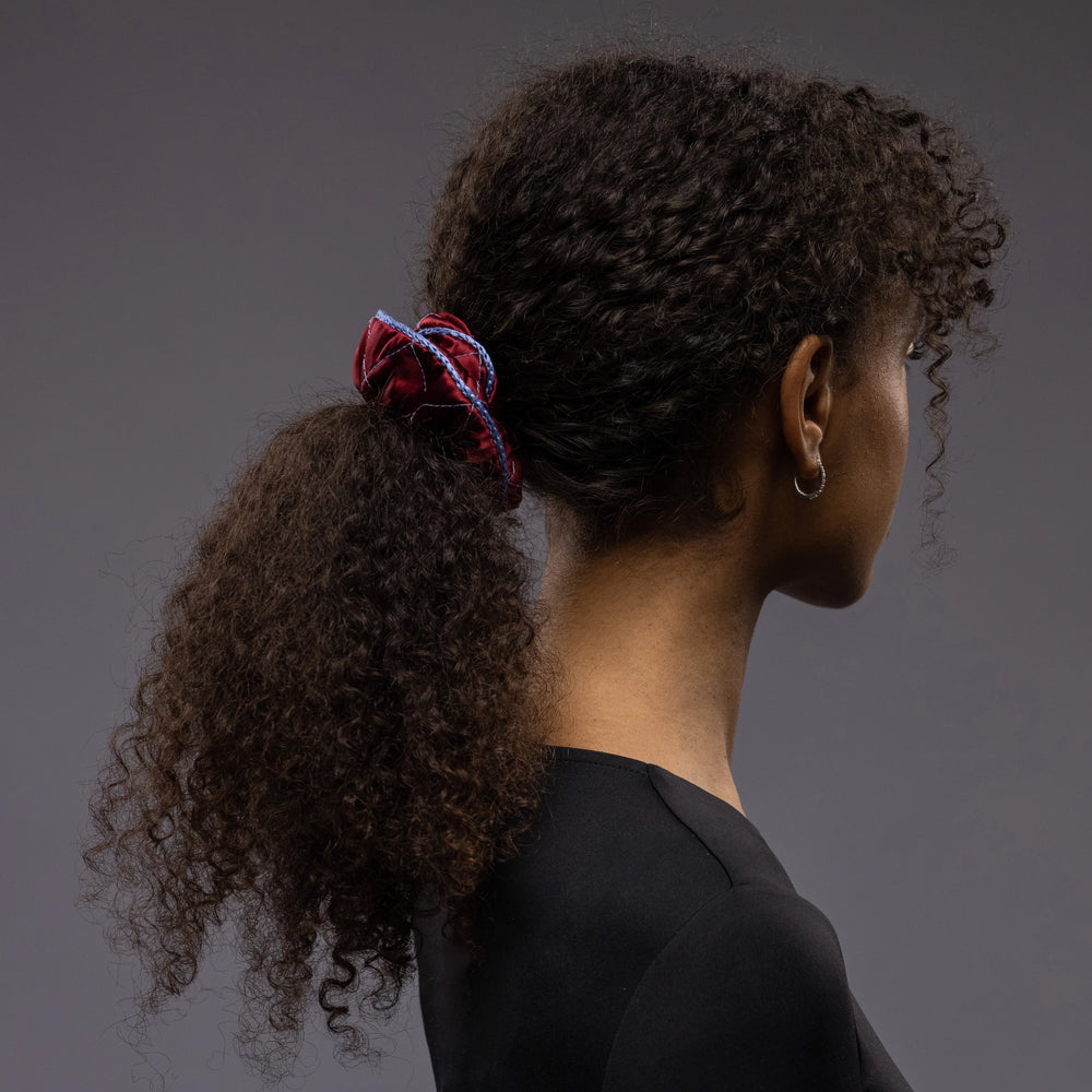 An African woman, view from the side, with stylish ponytail is wearing Malkiele designer burgundy quilted silk scrunchie, embellished with light blue silk knit ribbon, name Morning Passion