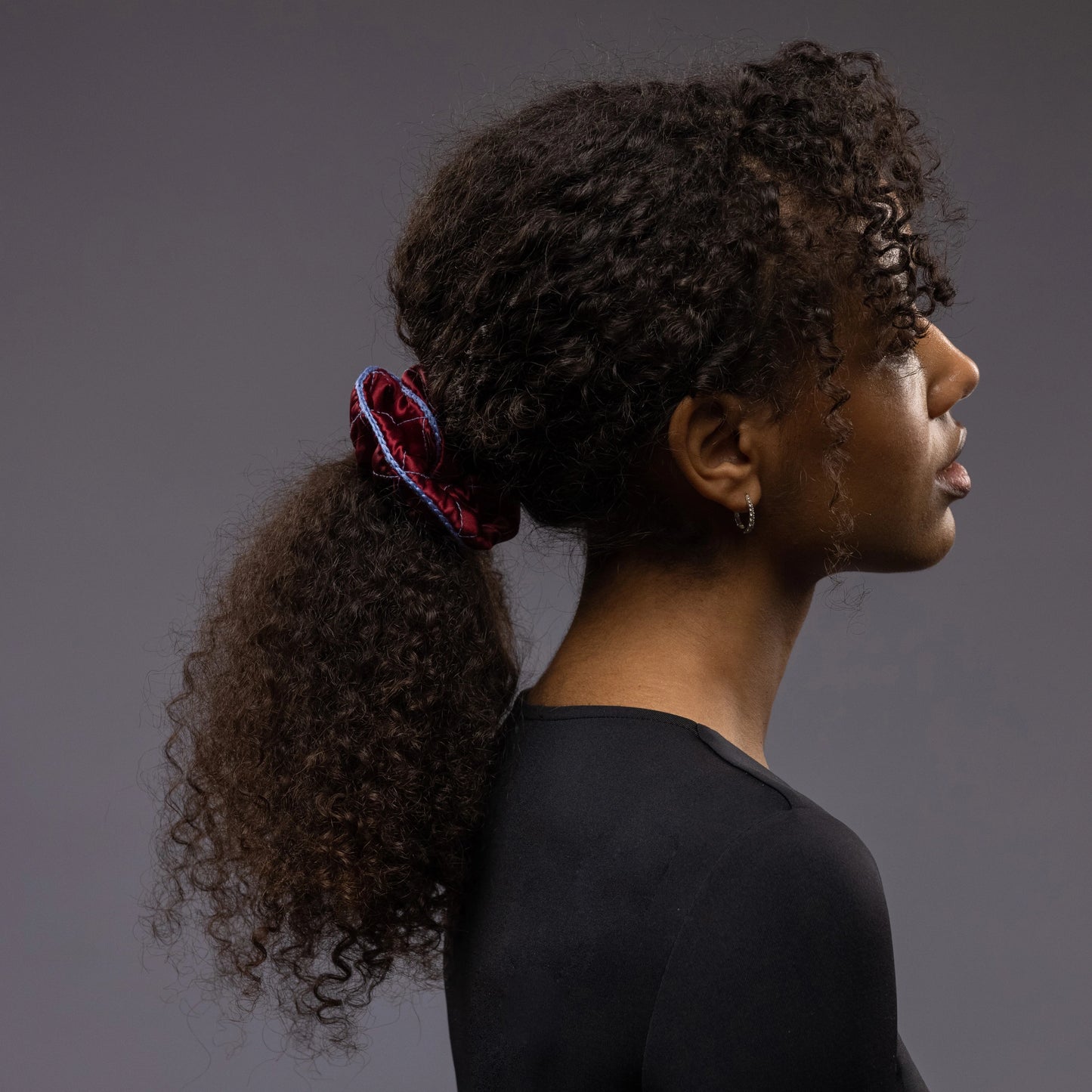 
                  
                    An African woman, view from the side, with stylish ponytail is wearing Malkiele designer burgundy quilted silk scrunchie, embellished with light blue silk knit ribbon, name Morning Passion
                  
                