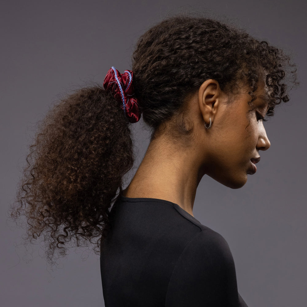 
                  
                    An African woman, view from the side, with stylish ponytail is wearing Malkiele designer burgundy quilted silk scrunchie, embellished with light blue silk knit ribbon, name Morning Passion
                  
                