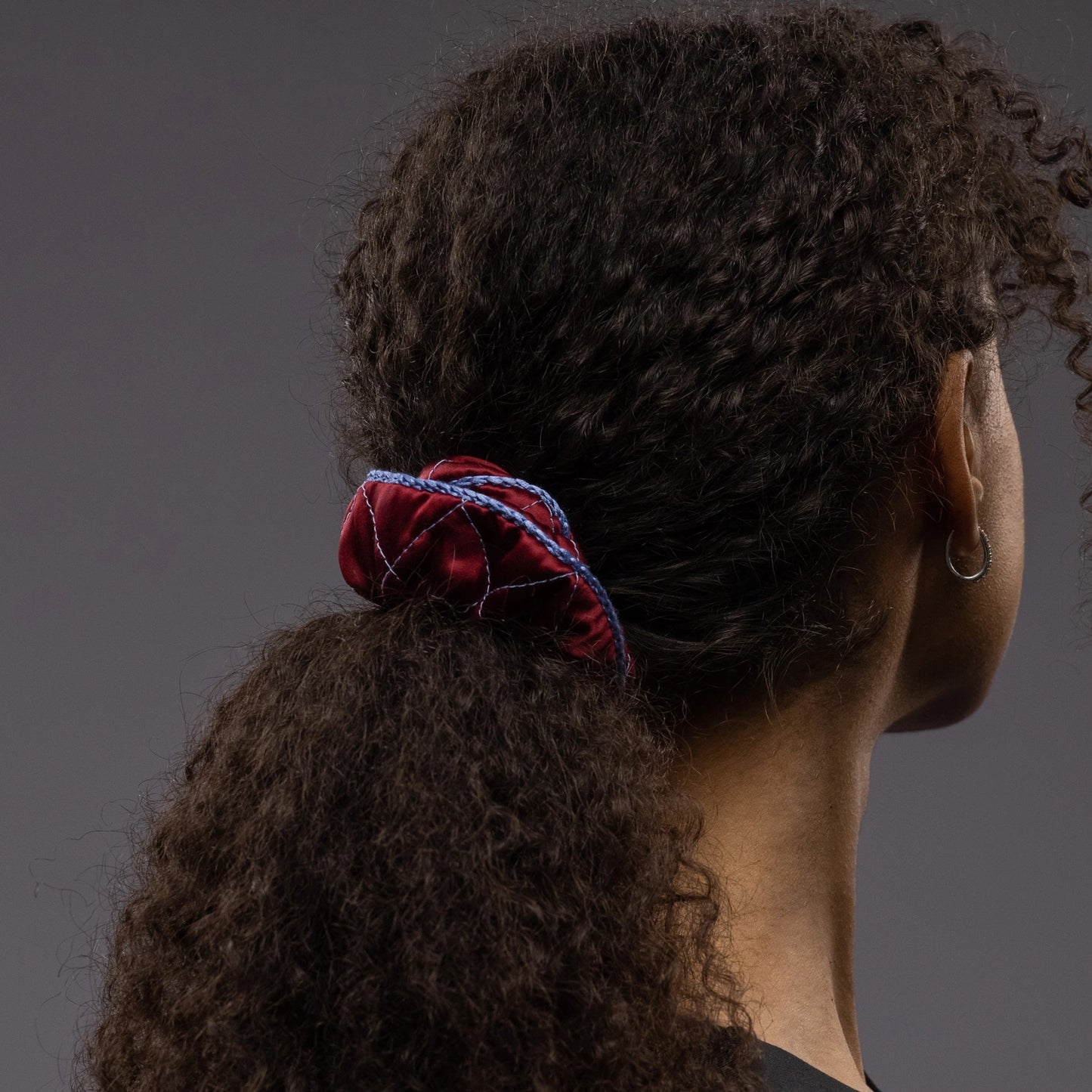 
                  
                    An African woman, view from the back, with stylish ponytail is wearing Malkiele designer burgundy quilted silk scrunchie, embellished with light blue silk knit ribbon, name Morning Passion
                  
                