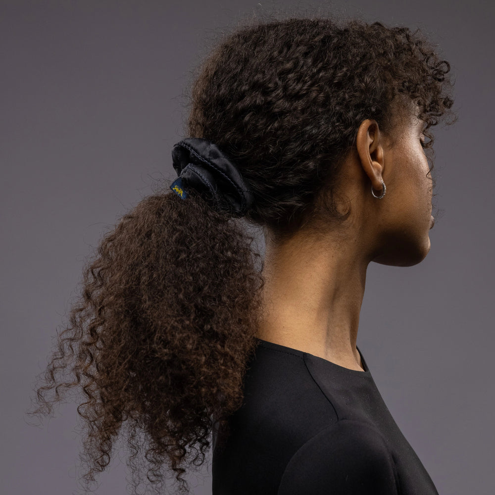 
                  
                    An African woman, view from the side, with long and stylish ponytail is wearing Malkiele designer black quilted silk scrunchie, embellished with black silk knit ribbon, name Morning Passion
                  
                