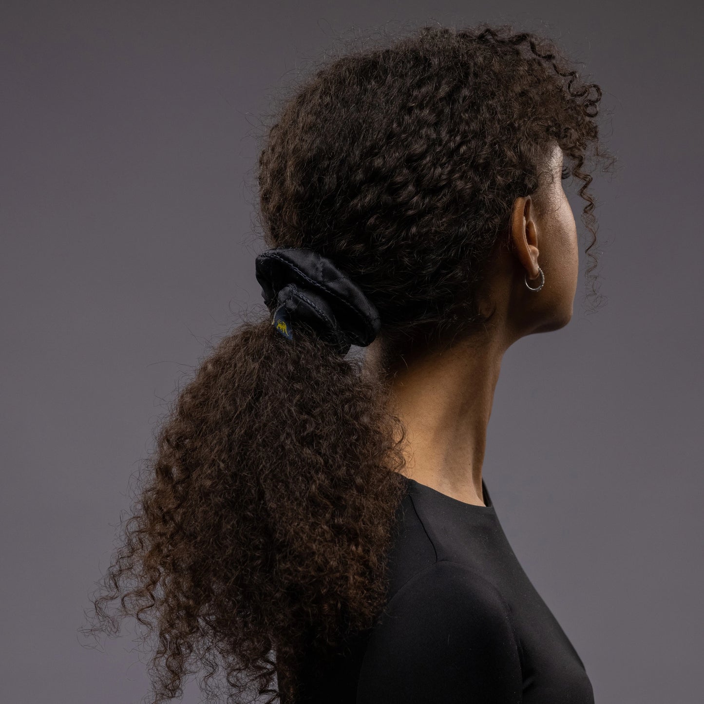 
                  
                    A dark-haired woman, view from the back, with long and stylish ponytail is wearing Malkiele designer black quilted silk scrunchie, embellished with black silk knit ribbon, name Morning Passion
                  
                