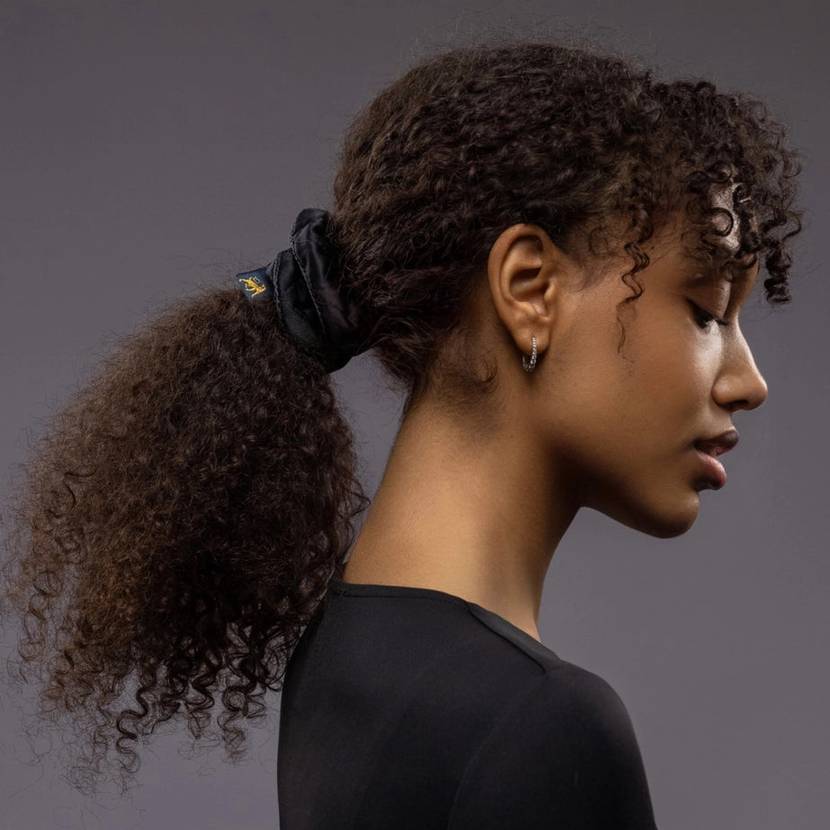 An African woman, view from the side, with long and stylish ponytail is wearing Malkiele designer black quilted silk scrunchie, embellished with black silk knit ribbon, name Morning Passion