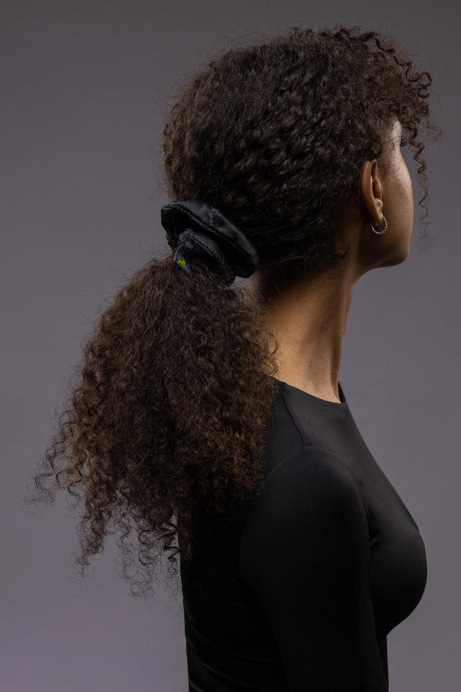 
                  
                    An African dark-haired woman, view from the back, with long and stylish ponytail is wearing Malkiele designer black quilted silk scrunchie, embellished with black silk knit ribbon, name Morning Passion
                  
                