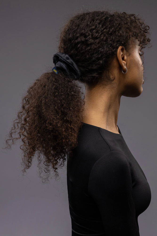 
                  
                    An African dark-haired woman, view from the side, with long and stylish ponytail is wearing Malkiele designer black quilted silk scrunchie, embellished with black silk knit ribbon, name Morning Passion
                  
                