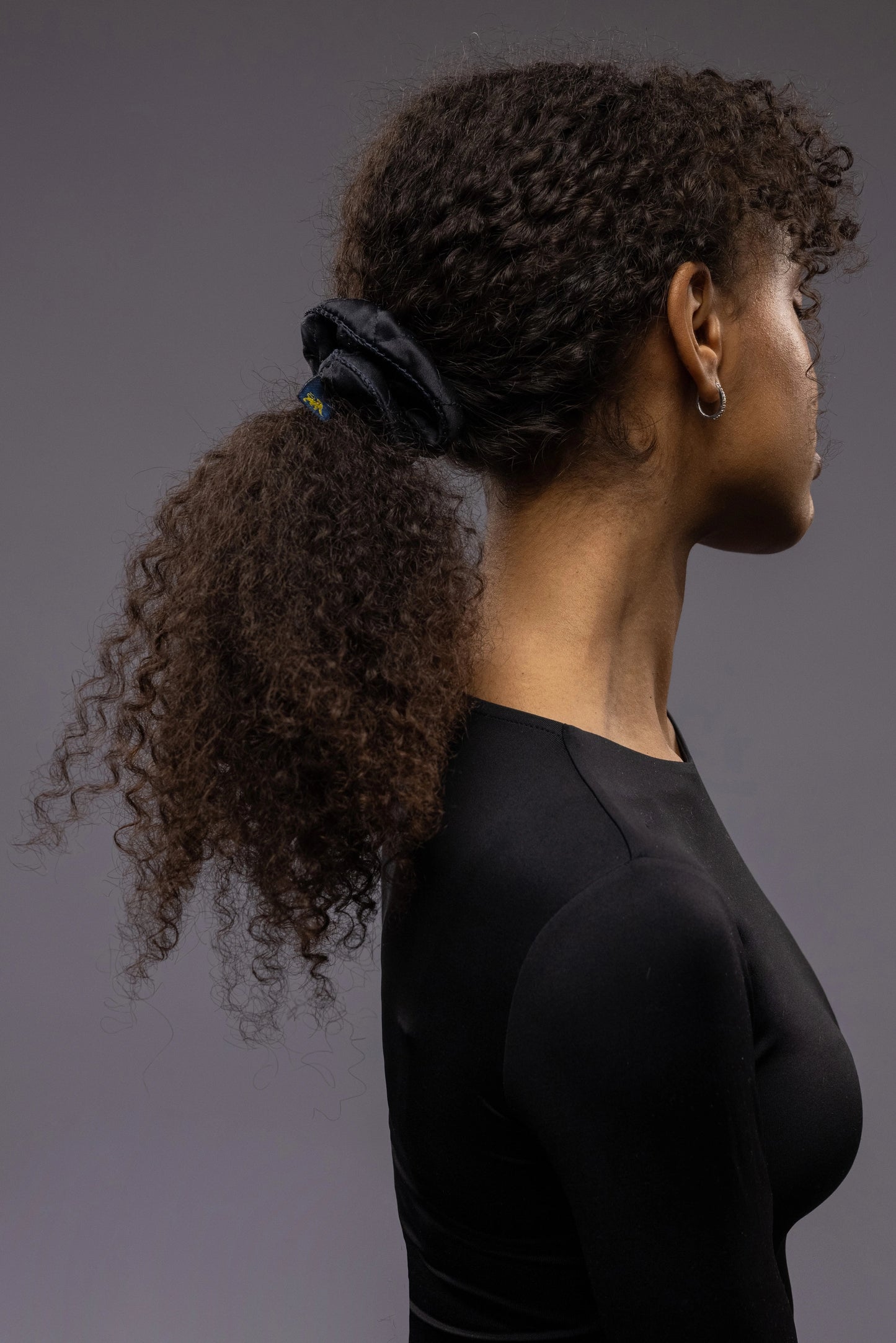 
                  
                    An African dark-haired woman, view from the side, with long and stylish ponytail is wearing Malkiele designer black quilted silk scrunchie, embellished with black silk knit ribbon, name Morning Passion
                  
                