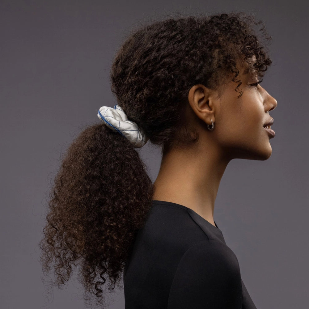 
                  
                    An African smiling woman, view from the side, half face showing, with a stylish ponytail is wearing Malkiele designer white quilted silk scrunchie, embellished with blue silk knit ribbon, name Morning Passion
                  
                