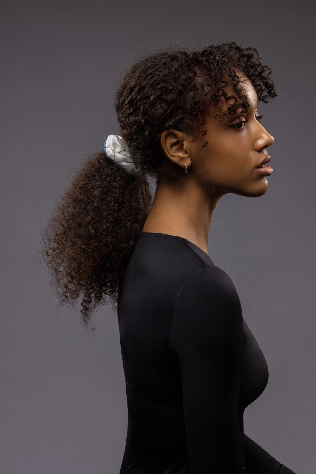 
                  
                    An African dark-haired woman, half face view from the side, with a stylish ponytail is wearing Malkiele designer white quilted silk scrunchie, embellished with blue silk knit ribbon, name Morning Passion
                  
                