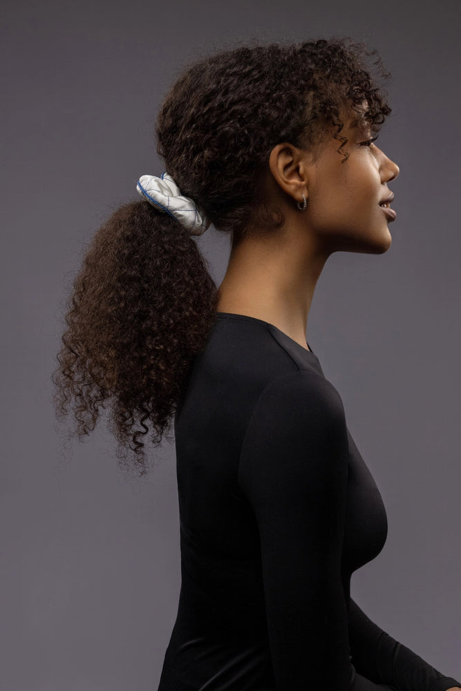 
                  
                    An African smiling woman, half face view from the side, with a stylish ponytail is wearing Malkiele designer white quilted silk scrunchie, embellished with blue silk knit ribbon, name Morning Passion
                  
                