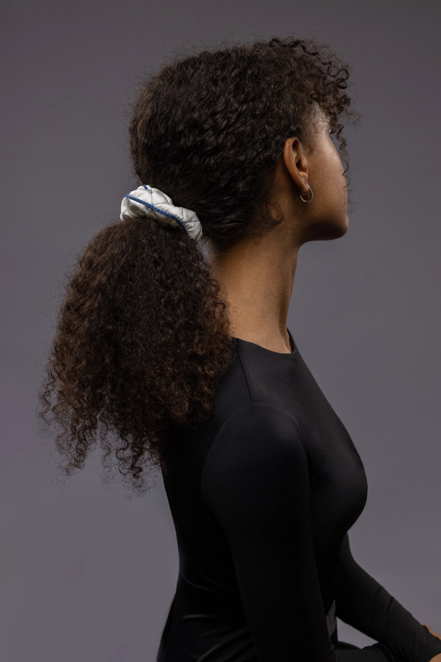 
                  
                    An African dark-haired woman, view from the back, with a stylish ponytail is wearing Malkiele designer white quilted silk scrunchie, embellished with blue silk knit ribbon, name Morning Passion
                  
                