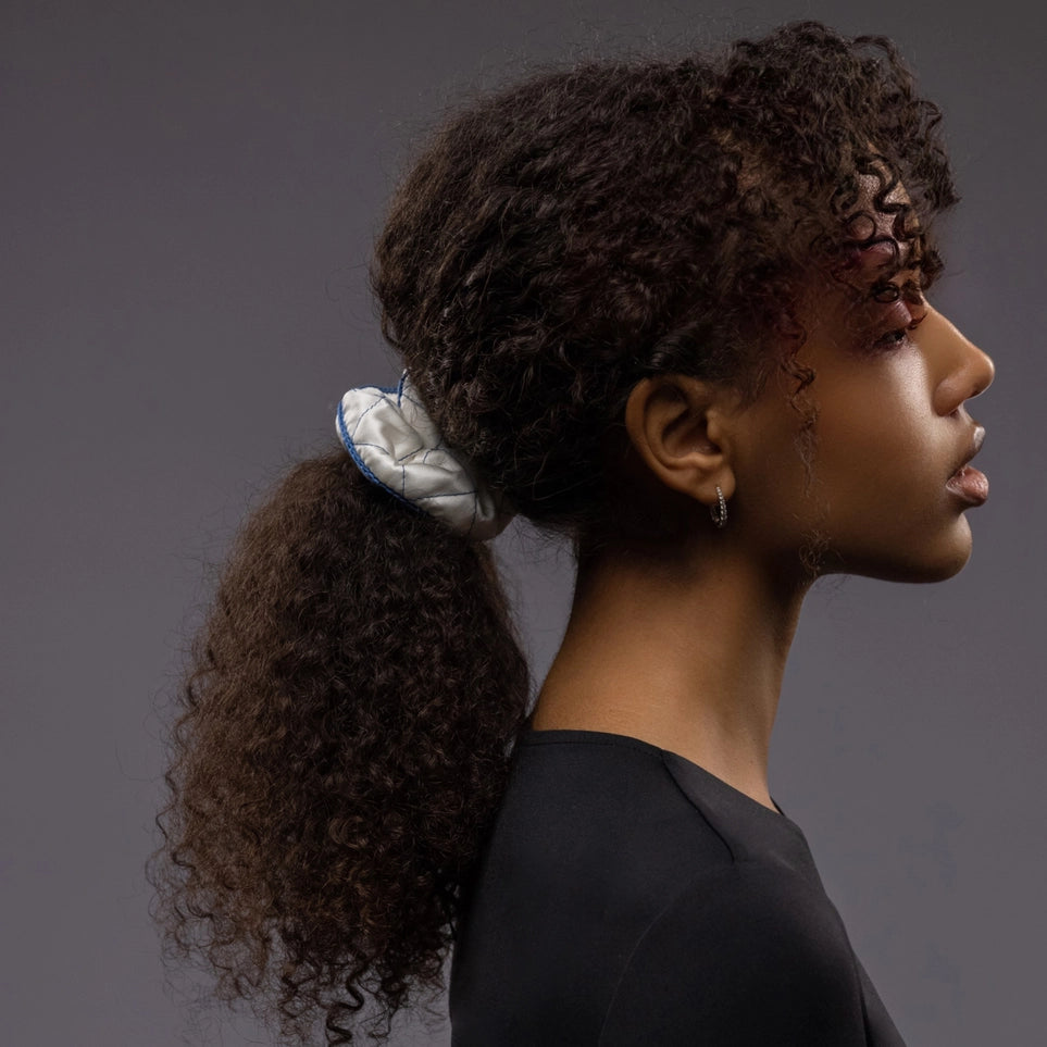 
                  
                    An African dark-haired woman, view from the side, half face showing, with a stylish ponytail is wearing Malkiele designer white quilted silk scrunchie, embellished with blue silk knit ribbon, name Morning Passion
                  
                