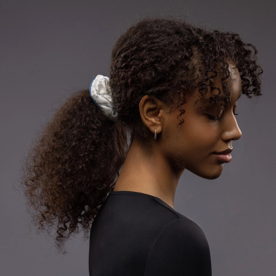 
                  
                    An African smiling woman, view from the side, with a stylish ponytail is wearing Malkiele designer white quilted silk scrunchie, embellished with blue silk knit ribbon, name Morning Passion
                  
                