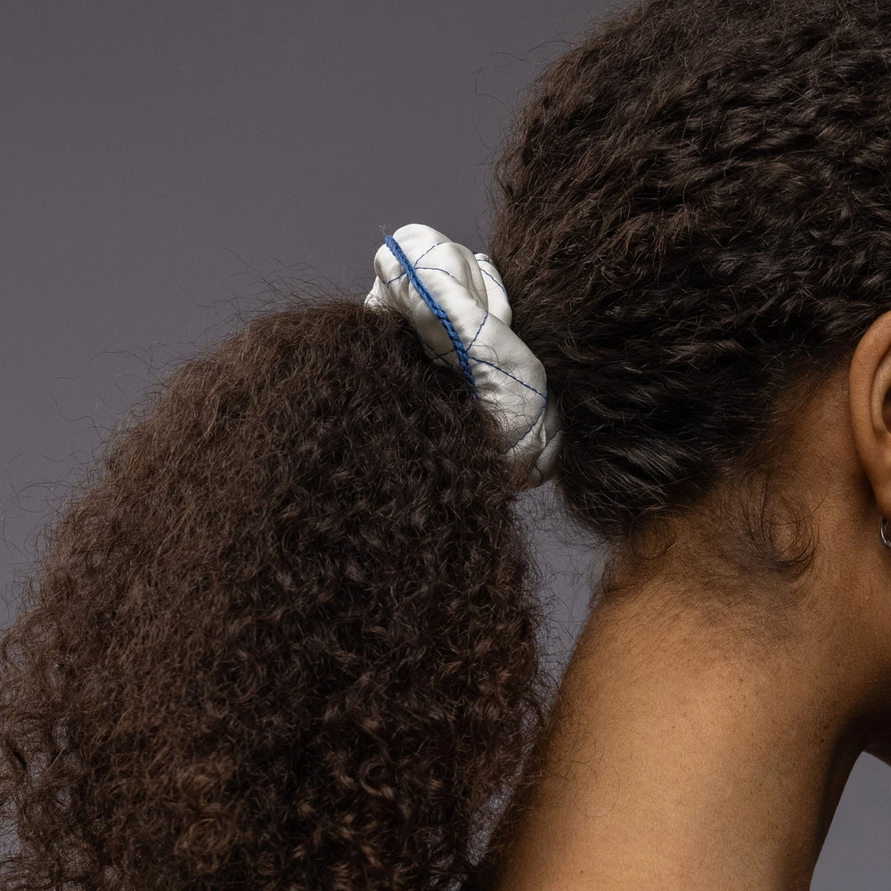 
                  
                    An African dark-haired woman, view from the side, details, with a stylish ponytail is wearing Malkiele designer white quilted silk scrunchie, embellished with blue silk knit ribbon, name Morning Passion
                  
                