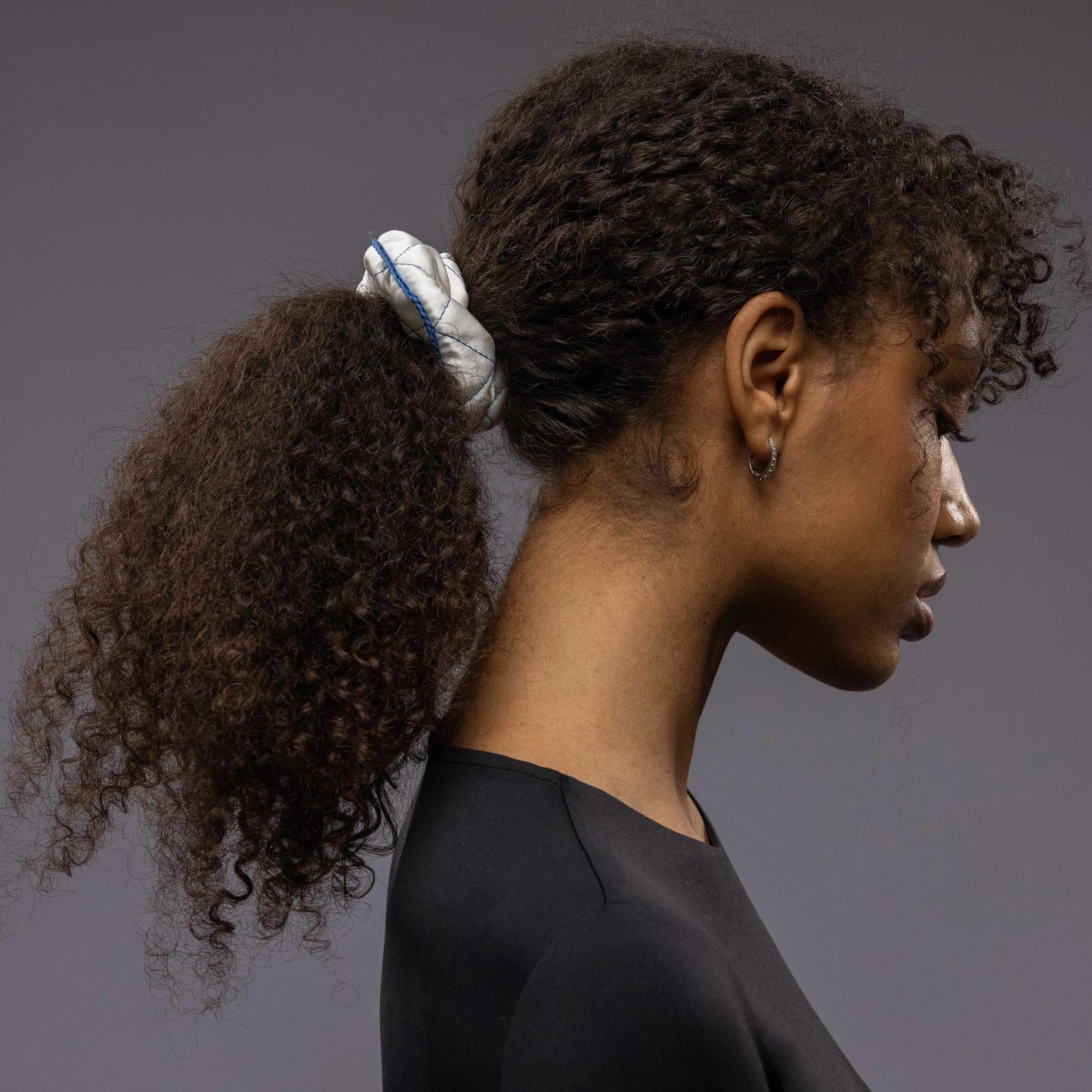 
                  
                    An African dark-haired woman, view from the side, half face showing, with a stylish ponytail is wearing Malkiele designer white quilted silk scrunchie, embellished with blue silk knit ribbon, name Morning Passion
                  
                