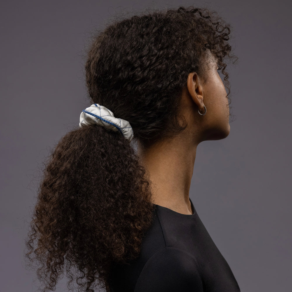 An African dark-haired woman, view from the side, with a stylish ponytail is wearing Malkiele designer white quilted silk scrunchie, embellished with blue silk knit ribbon, name Morning Passion