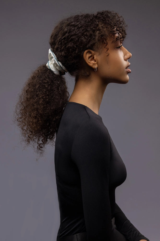 
                  
                    An African dark-haired woman, view from the side, half face showing, with a stylish ponytail is wearing Malkiele designer white quilted silk scrunchie, embellished with green silk knit ribbon, name Morning Passion
                  
                