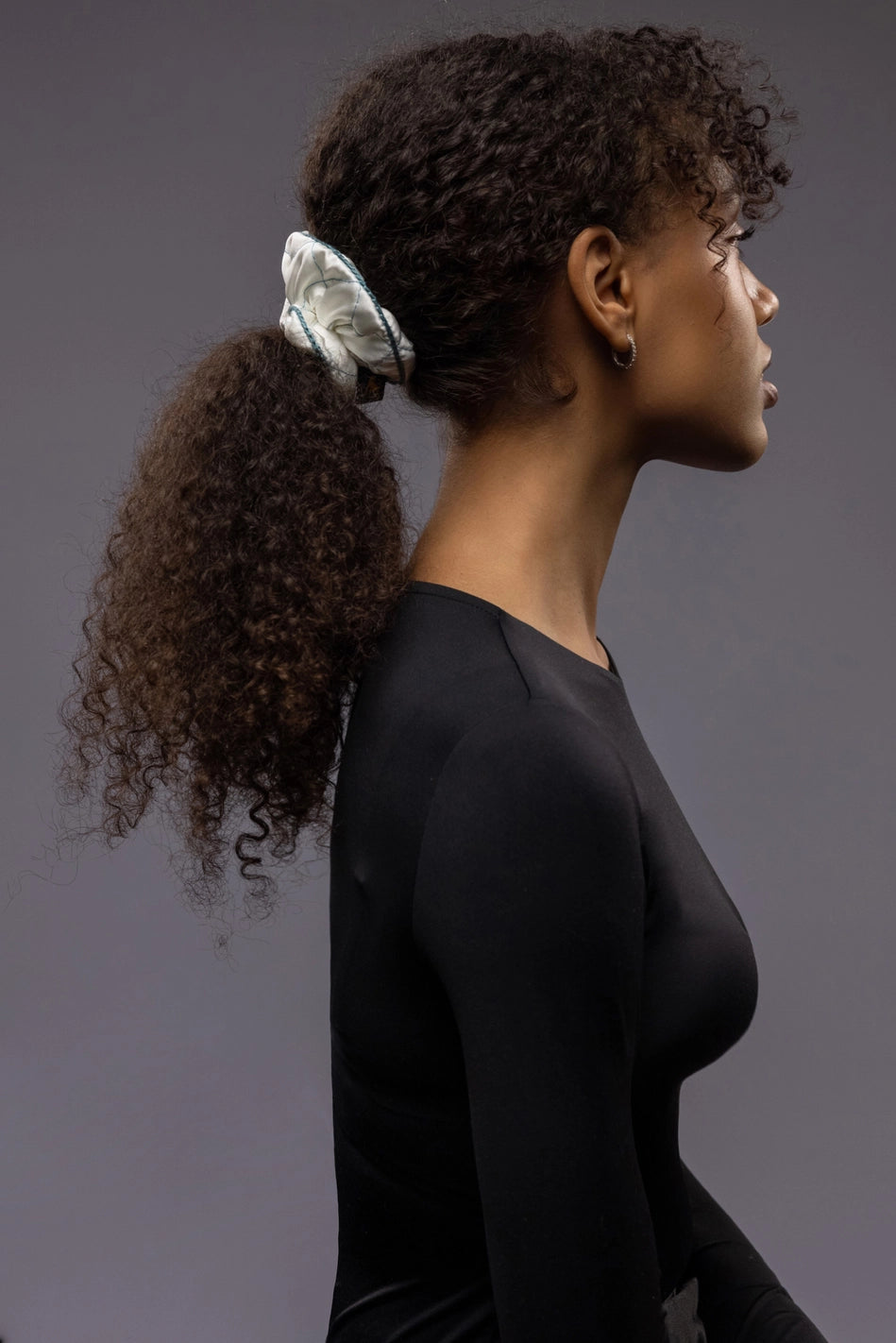 
                  
                    An African dark-haired woman, view from the side, with a stylish ponytail is wearing Malkiele designer white quilted silk scrunchie, embellished with green silk knit ribbon, name Morning Passion
                  
                