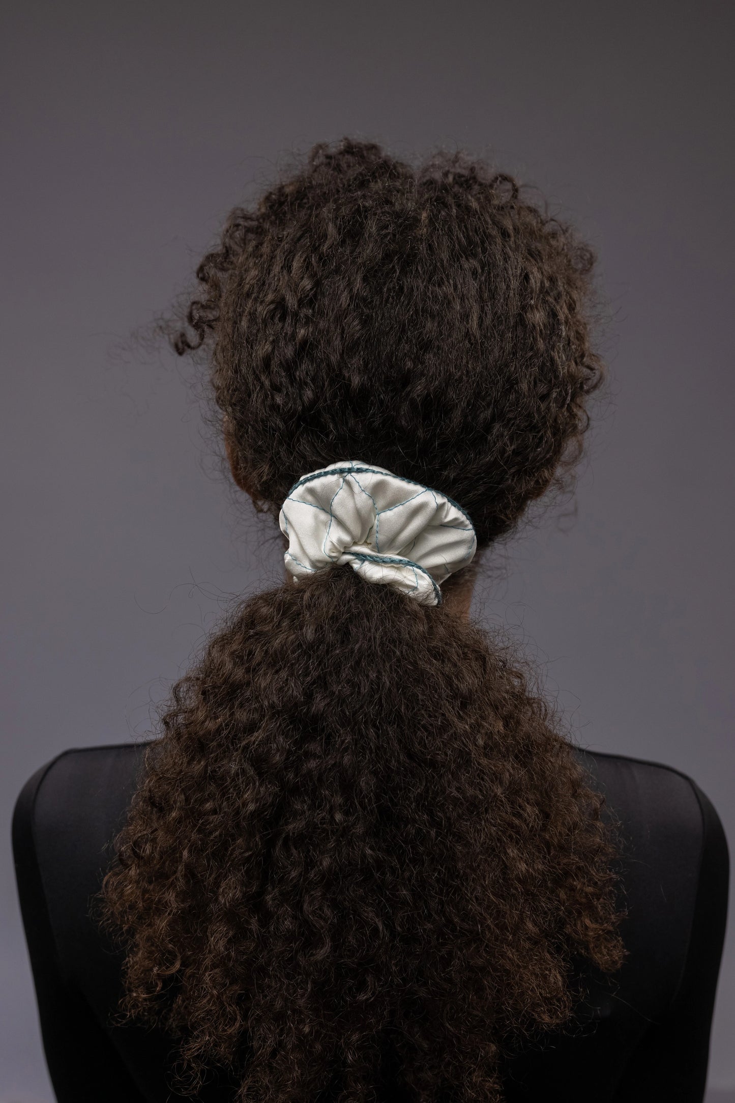 
                  
                    An African dark-haired woman, view from the back, with a stylish ponytail is wearing Malkiele designer white quilted silk scrunchie, embellished with green silk knit ribbon, name Morning Passion
                  
                