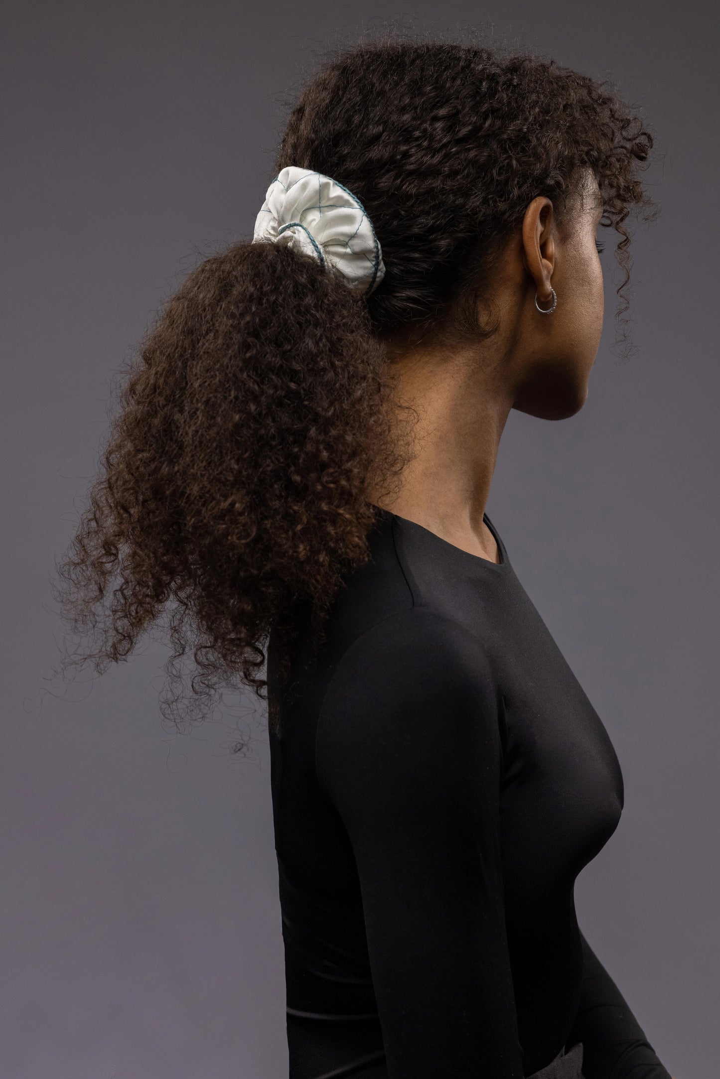 
                  
                    An African dark-haired woman, view from the side, with a stylish ponytail is wearing Malkiele designer white quilted silk scrunchie, embellished with green silk knit ribbon, name Morning Passion
                  
                