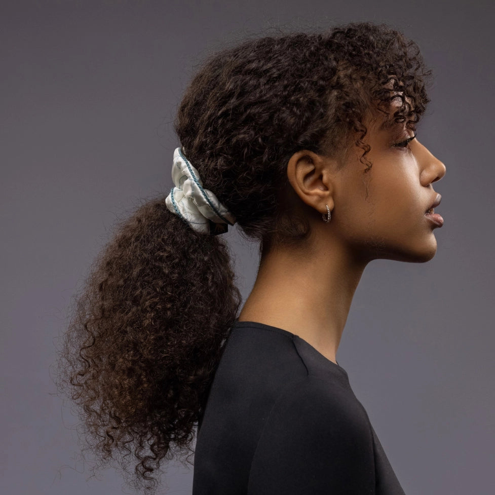 An African dark-haired woman, view from the side, half face showing, with a stylish ponytail is wearing Malkiele designer white quilted silk scrunchie, embellished with green silk knit ribbon, name Morning Passion
