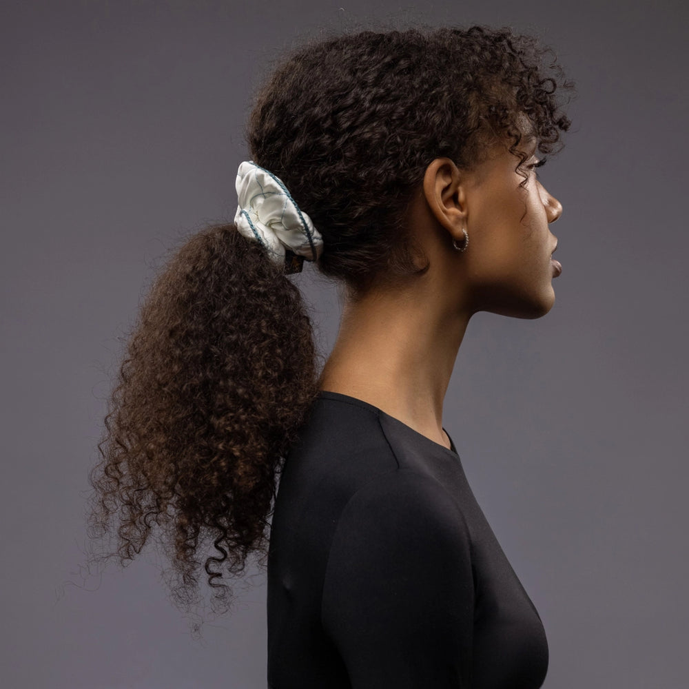 
                  
                    An African dark-haired woman, view from the side, with a stylish ponytail is wearing Malkiele designer white quilted silk scrunchie, embellished with green silk knit ribbon, name Morning Passion
                  
                