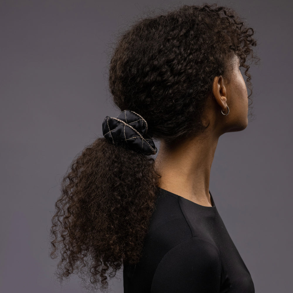 An African dark-haired woman, view from the back, with long and stylish ponytail is wearing Malkiele designer black quilted silk scrunchie, embellished with beige silk knit ribbon, name Morning Passion