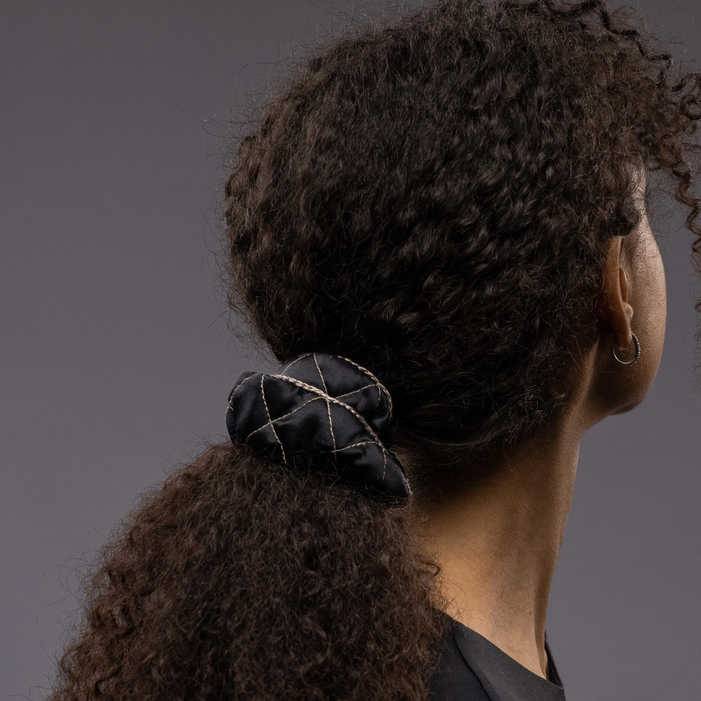 
                  
                    An African dark-haired woman, view from the back, with a stylish ponytail is wearing Malkiele designer black quilted silk scrunchie, embellished with beige silk knit ribbon, name Morning Passion
                  
                