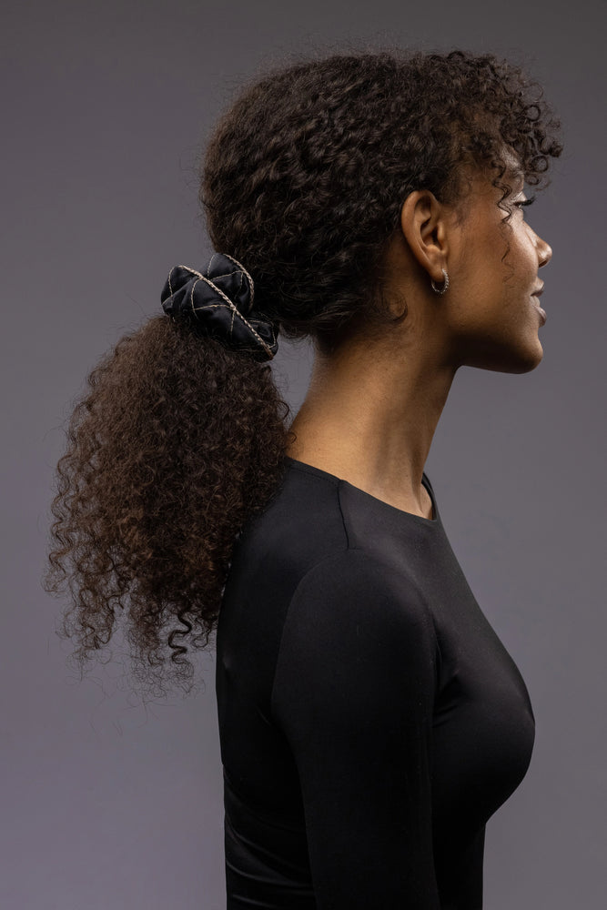 
                  
                    An African smiling woman, view from the side, with stylish ponytail is wearing Malkiele designer black quilted silk scrunchie, embellished with beige silk knit ribbon, name Morning Passion
                  
                