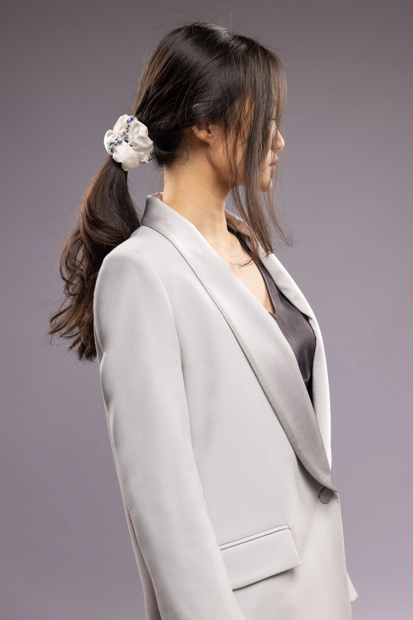 
                  
                    A dark-haired woman in grey jacket, side view, with a stylish ponytail is wearing Malkiele designer white silk scrunchie, embellished with blue pearls, name Elizabeth
                  
                