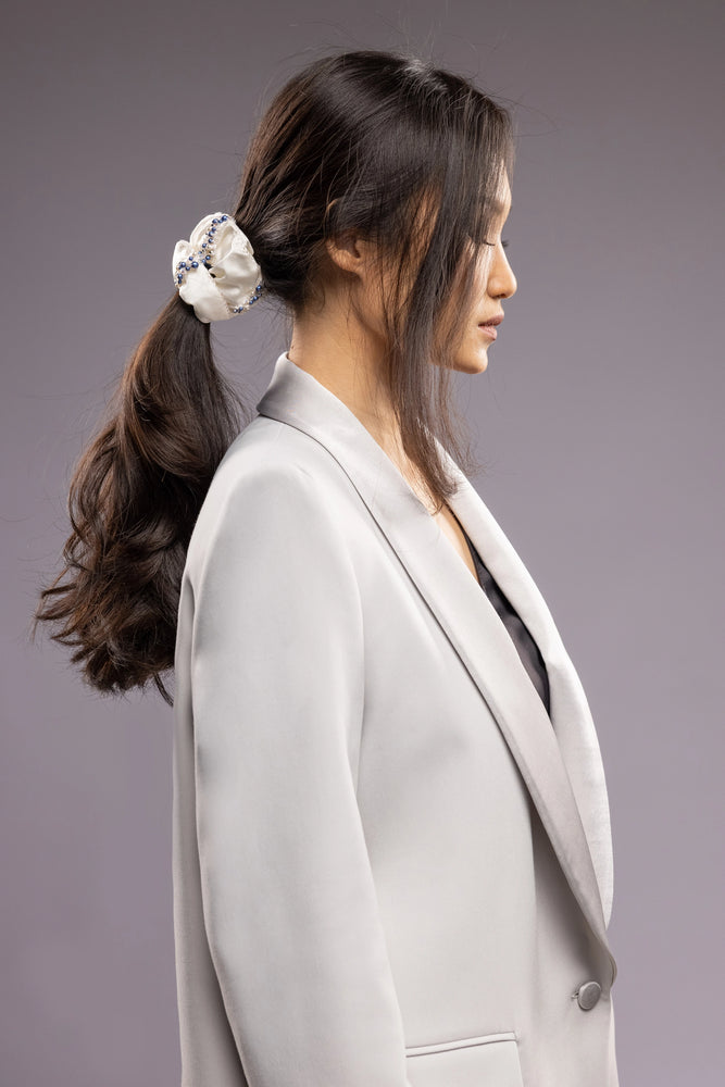 
                  
                    A dark-haired woman in grey jacket, view from the side, with a stylish ponytail is wearing Malkiele designer white silk scrunchie, embellished with blue pearls, name Elizabeth
                  
                