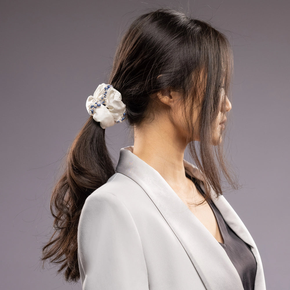 
                  
                    A dark-haired woman in grey jacket, view from the side, with a stylish ponytail is wearing Malkiele designer white silk scrunchie, embellished with blue pearls, name Elizabeth
                  
                