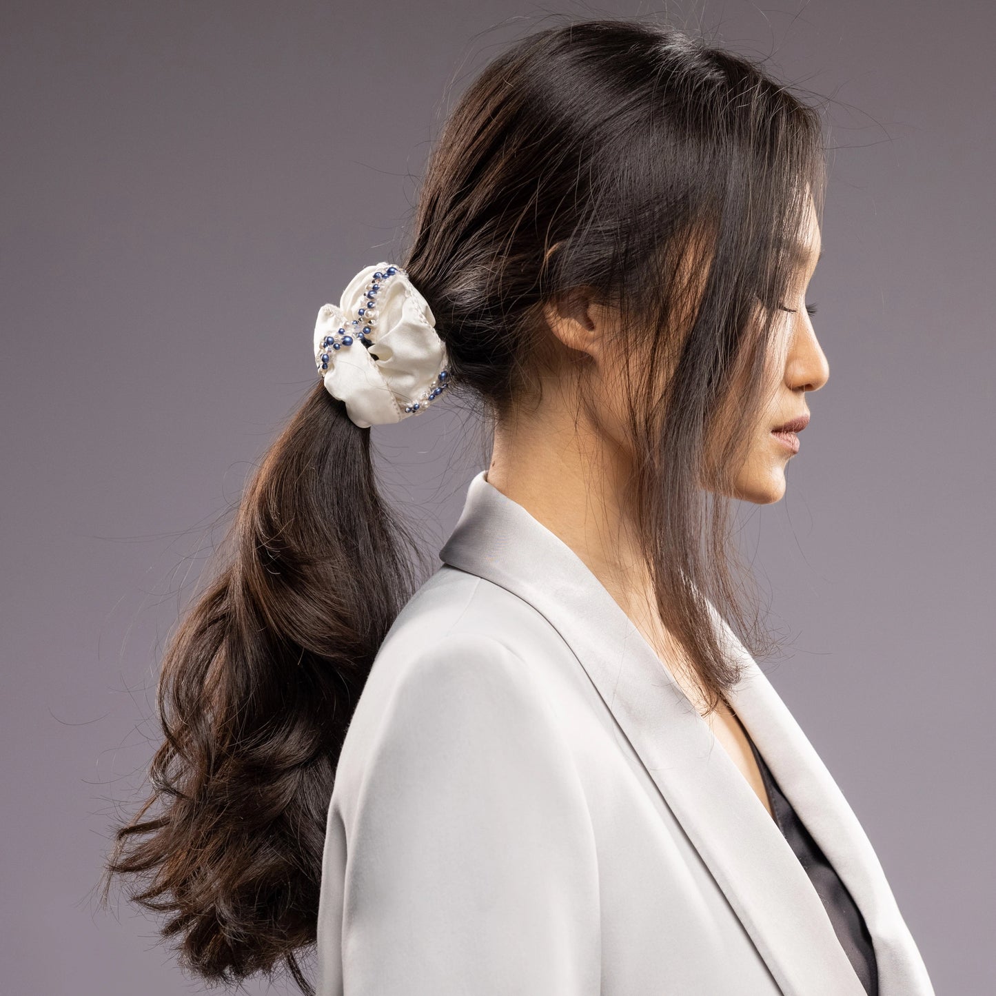 
                  
                    A dark-haired woman in grey jacket, view from the side, with a stylish ponytail is wearing Malkiele designer white silk scrunchie, embellished with blue pearls, name Elizabeth
                  
                